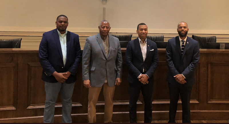 Four Black men in suits standing in front of a wooden paneled desk.