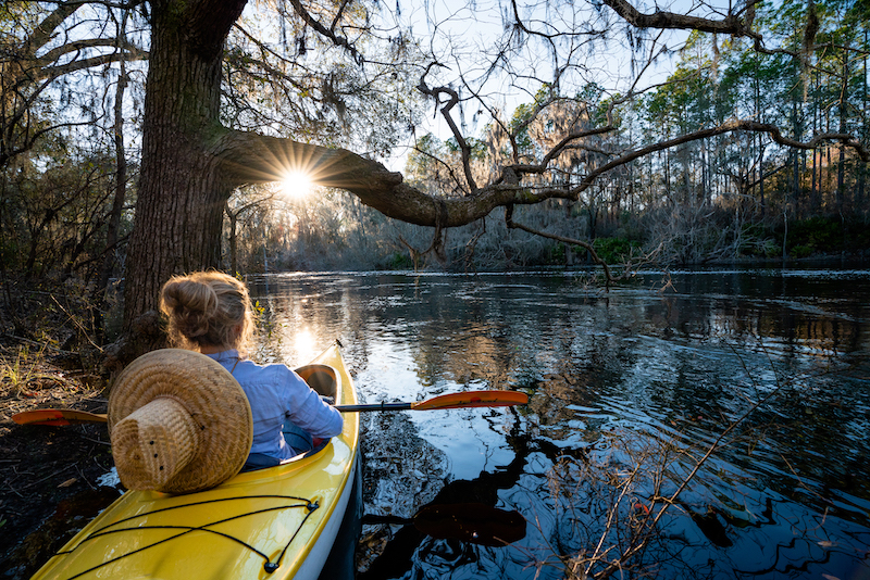 A woman in a yellow kayak paddling through Florida waterway with the sun setting through tree branches.